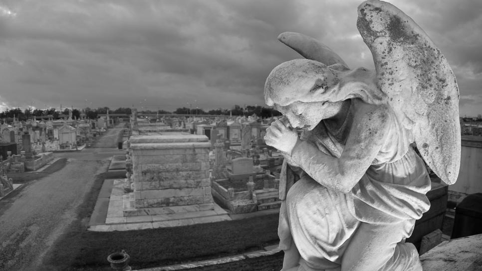 A praying statue kneels on top of a tomb in New Orleans under moody skies.  High point of view to contain the cemetery landscape in the background.